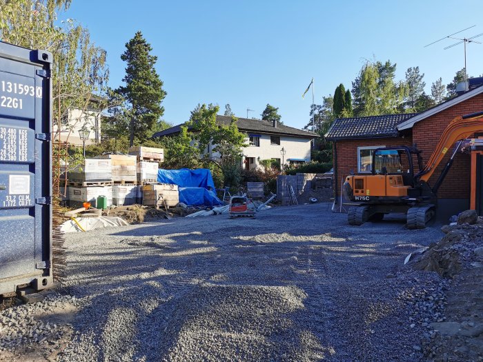 Byggarbetsplats med utspridd makadam, en grävmaskin, material och hus i bakgrunden.