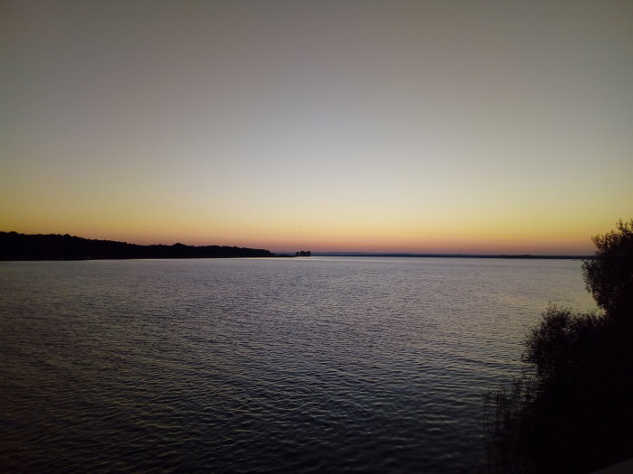 Vy över en stilla sjö vid solnedgång med silhuett av träd och en himmel i avtonade färger.