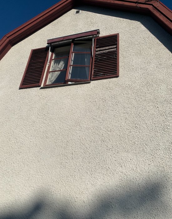 Fönsterlucka på grovkornig putsad fasad med öppna röda persienner och blå himmel ovanför.