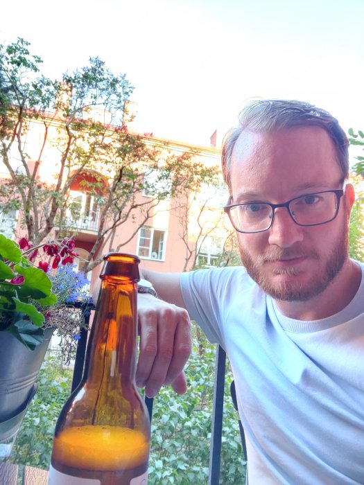 Man på balkong med ölflaska i hand, växter och röd byggnad i bakgrunden.