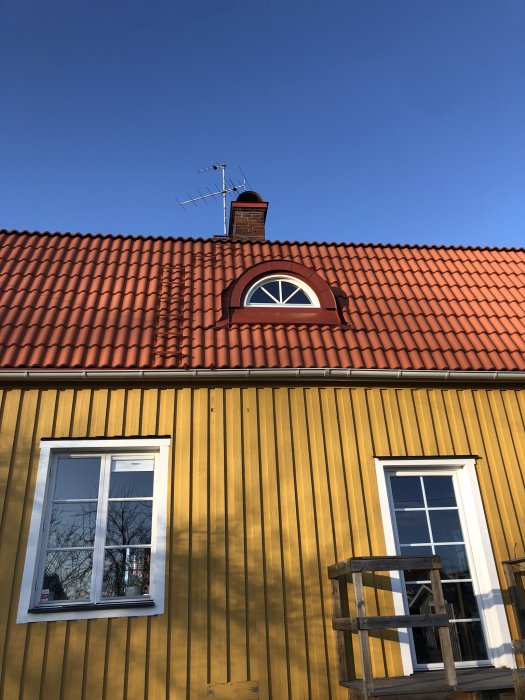 Välbuilt takkupa med halvrunt fönster installerat på ett rött tegeltak med gulmålad träfasad och en skorsten i bakgrunden.