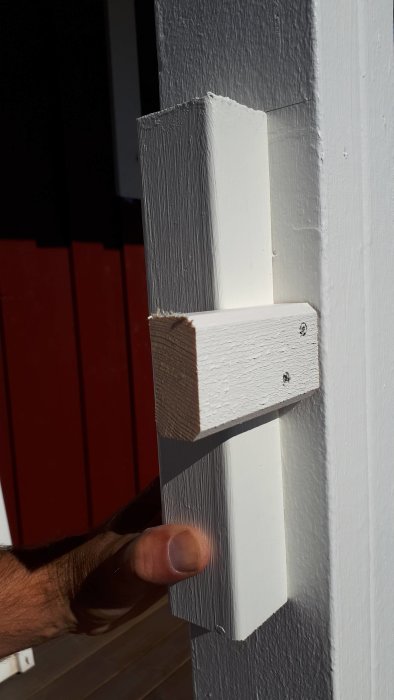 Lockläkt av trä fäst mot en vit stolpe med skruvar, hand som håller träbiten, balkong i bakgrunden.
