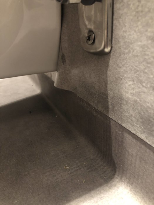 Delvis lyft plastmatta i hörnet vid en vägghängd toalett, tecken på möjlig fuktskada eller felaktig installation.