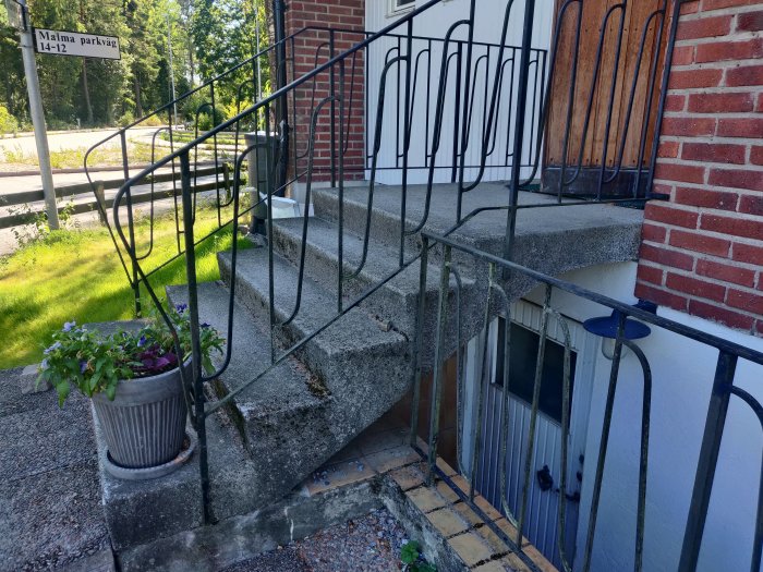 Slitna betongtrappor till ett hus med svart smidesräcke och en blomkruka vid entrén.