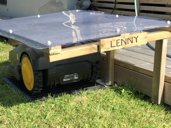 Robotgräsklippare vid laddstation under hemmagjort träskydd märkt med "LENNY".