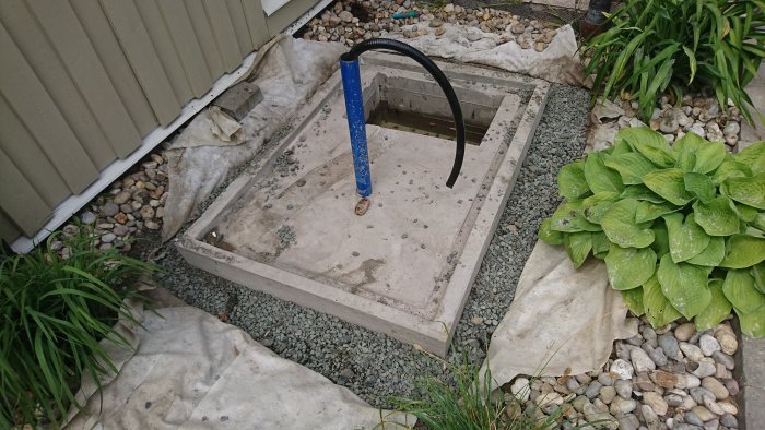 Betongfundament för pump med armeringsnät, blått rör och omgivande trädgårdsgrus och växter.
