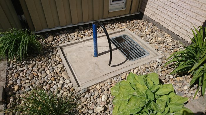 Betongfundament med armeringsnät, blå rörstång och svarta sladdar, omgivet av grus och växtlighet nära husvägg.