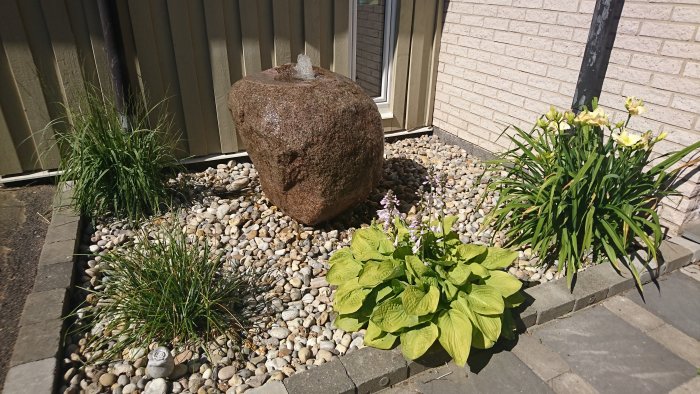 Trädgårdsdesign med stenfontän omgiven av småsten, gröna växter och blommor intill en byggnad.