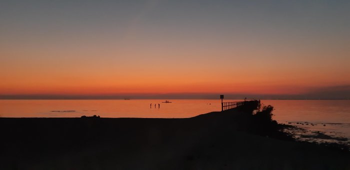 Solnedgång vid Barsebäckstrand med silhuetter av personer i vattnet och en brygga mot en orange himmel.