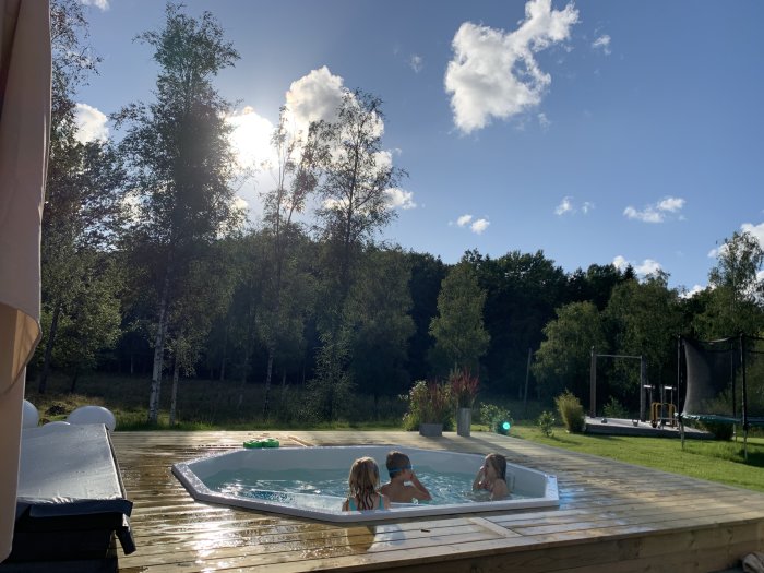 Barn badar i ett ovanmarkspool på en trädäck vid skogskant under solig himmel.