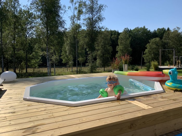 Barn i simglasögon flyter i pool på trädäck med trädgård i bakgrunden.