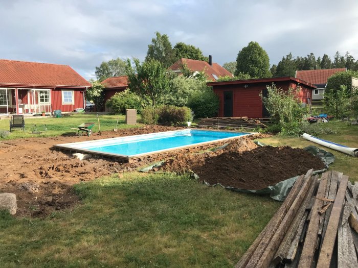 Renoveringsarbete vid poolområde med uppriven trädäck och synlig jord, gräshög och byggmaterial. Omgiven av grönska och röda hus.