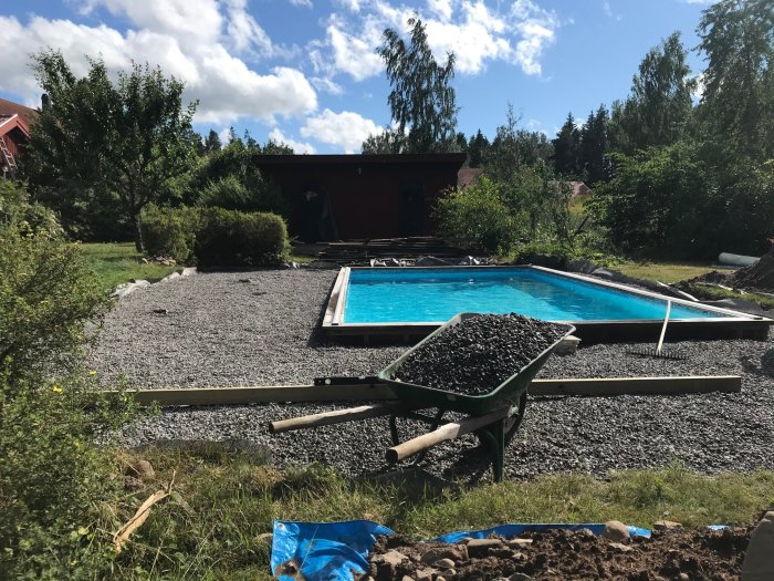 Renoveringsprojekt av trädäck vid pool med spridd grus, skottkärra med grus och ojämn gräsyta.