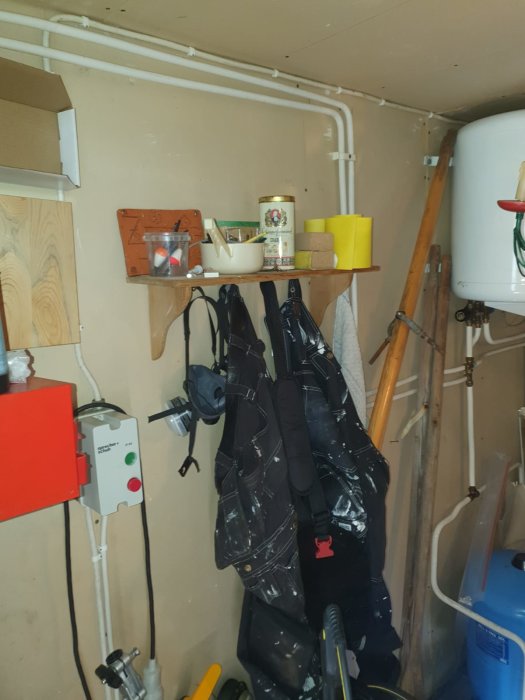 Inredning i förråd med synliga vita rör längs väggen, en varmvattenberedare och diverse föremål och verktyg på hyllor.
