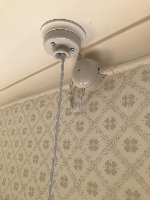 Utanpåliggande elinstallation med tvinnad kabel och porslinskopp på mönstrad tapet.