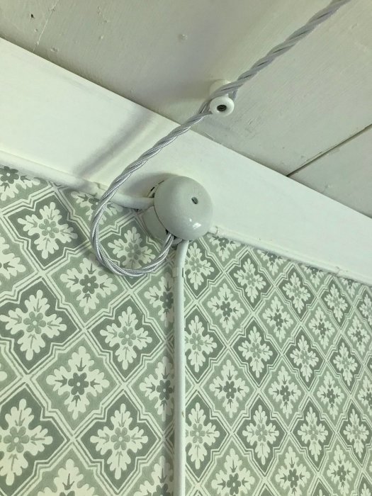 Utanpåliggande elinstallation med tvinnad kabel och porslinskopp längs mönstrad tapet och taklist.