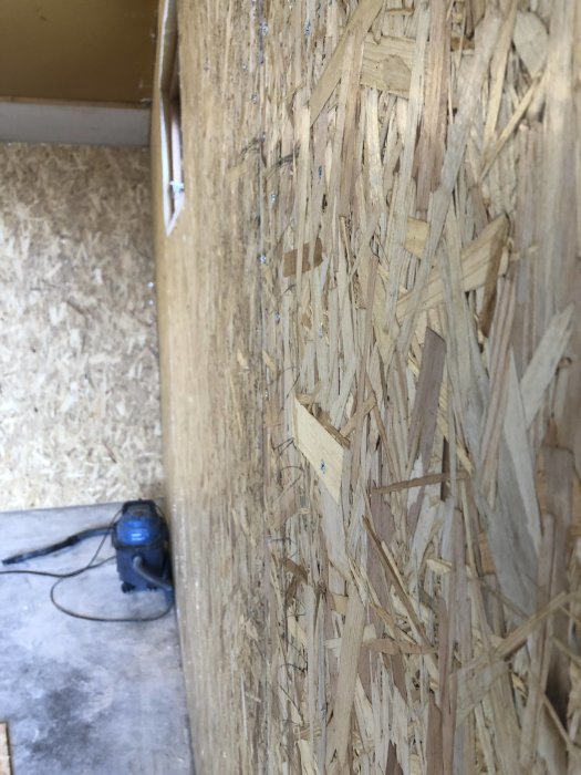 Tettbilde av vegg med OSB-plater i et verkstedområde, med støvsuger i bakgrunnen.