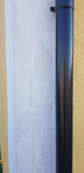 En vit husvägg med små sprickor och ett svart stuprör.