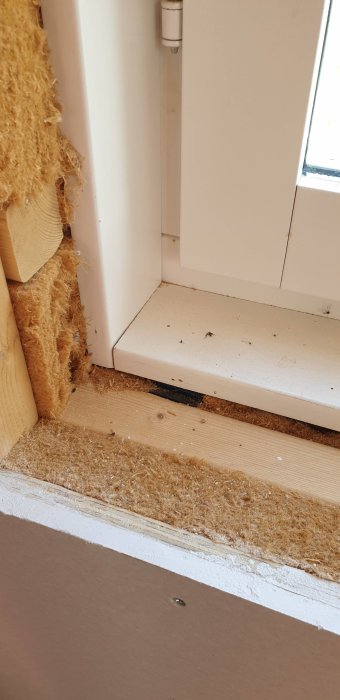 Detaljbild av en fönsterinstallation med synliga isoleringsskikt och en vit fönsterkarm utan fästelement för smygen.