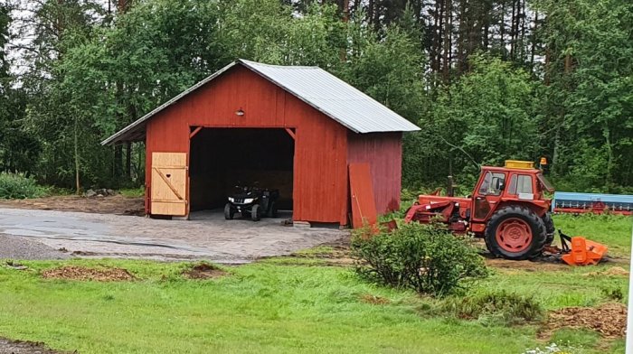 En röd lada med öppen port och padat grus framför, en ATV inuti och en röd traktor ute till höger.