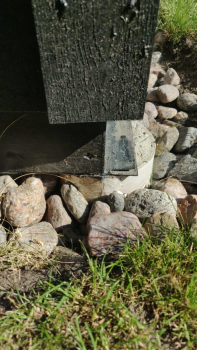 Träregel monterad på en betongplint omgiven av stenar och gräs, möjlig installationspunkt för plattjärn.