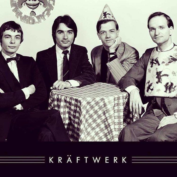 Svartvit bild av bandet Kraftwerk i kostymer, en med festhatt, sittande bakom ett bord med rutig duk.