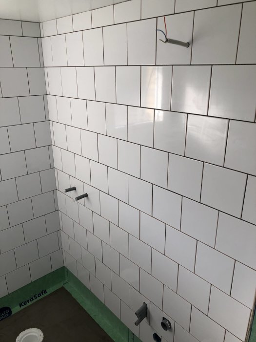 Nyinstallerade vita kakelplattor på vägg i badrum med synlig rörgenomföring och golv tätskikt.