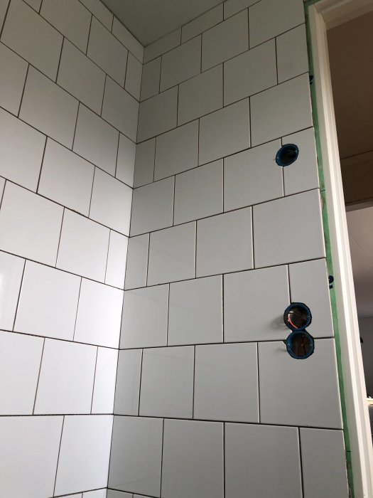Nyinstallerade vita kakelplattor på vägg med synliga hörn, eluttag och en ojämn fog ovanför en dörrkarm.