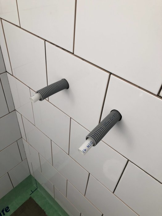 Nyligen kaklade badrumsväggar med två utstickande röröverdrag och små kakelbitar längst ner.