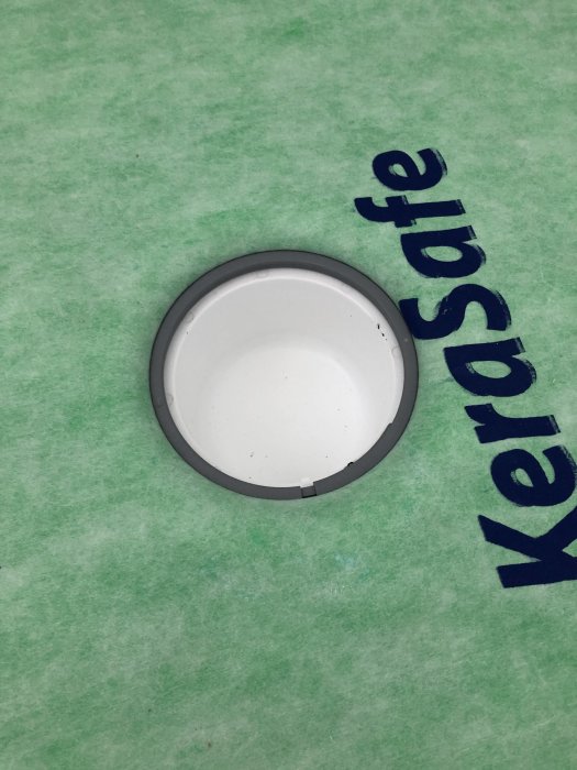 Klämring för rörtättning monterad i grön tätskiktsmatta, med texten "KerdiSafe" synlig, som del av badrumsrenovering.