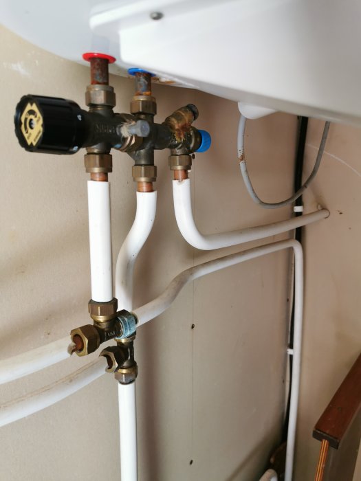 Rördragning med kopplingar och ventiler under vit varmvattenberedare mot brun vägg.