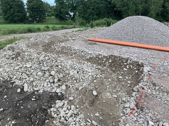 Uppgrävd mark med orange dagvattenrör och grushög, förberedd för dränering vid husgrund.