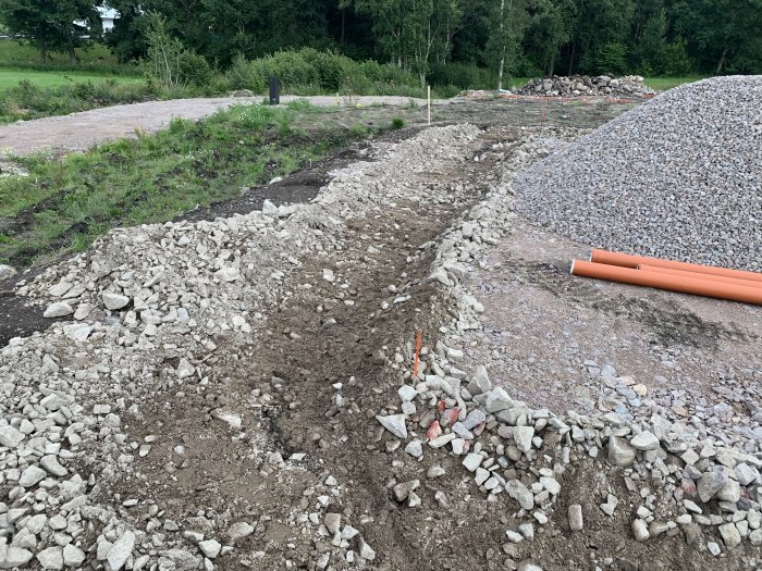 Uppgrävd grusig yta för dränering med orangea rör redo, vid ett husbygge.