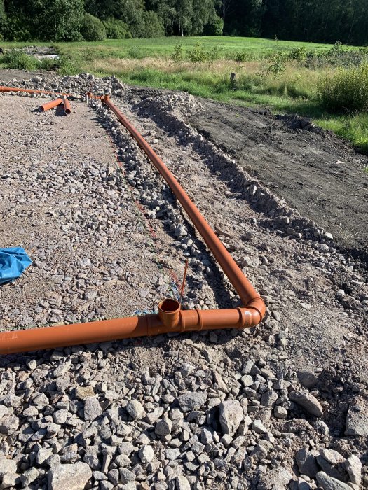 Nyinstallerat dagvattensystem med orange rör i en grävd ränna omringad av grus och jord i en trädgård.