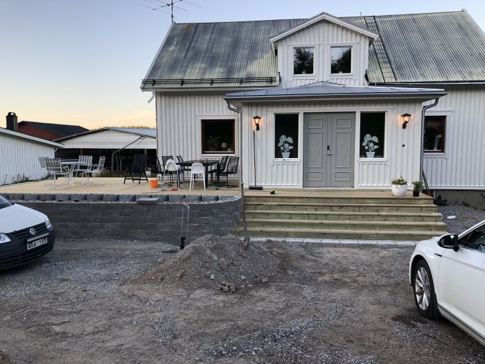 Nybyggd altan och mur framför ett vitt hus med dubbla dörrar och en utbyggnad, grus och påbörjad marksten till höger.