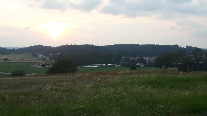 Solnedgång över ett lantligt landskap med hus och gårdar i förgrunden och träd i bakgrunden.