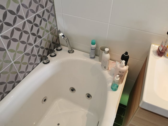 Ett bubbelbadkar intill ett badrumsskåp och vägg med färgglada kakelplattor, omgiven av badrumsprodukter.