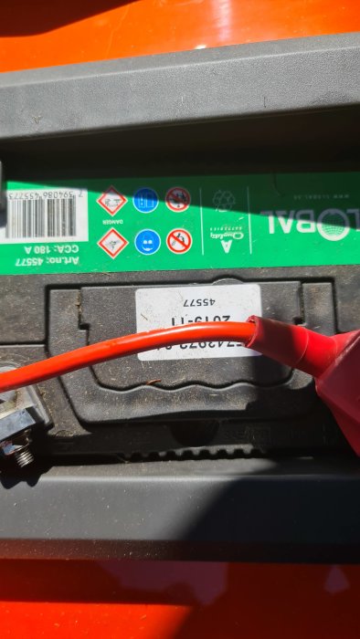 Röd batteriklämma ansluten till bilbatteris pluspol med säkerhets- och varningsikoner i bakgrunden.