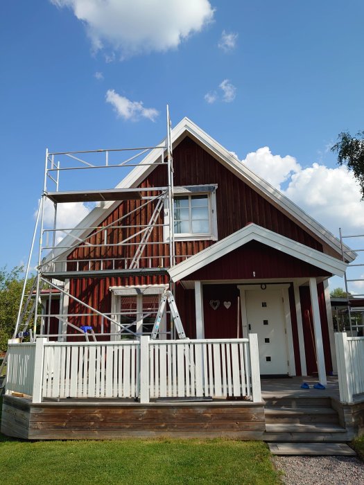 Rött hus med uppfälld byggställning och halvfärdigt arbete på taknocken under solig himmel.