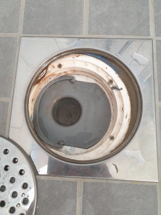 Öppnad golvbrunn i badrum med synligt smuts och avlagringar runt avloppshålet.