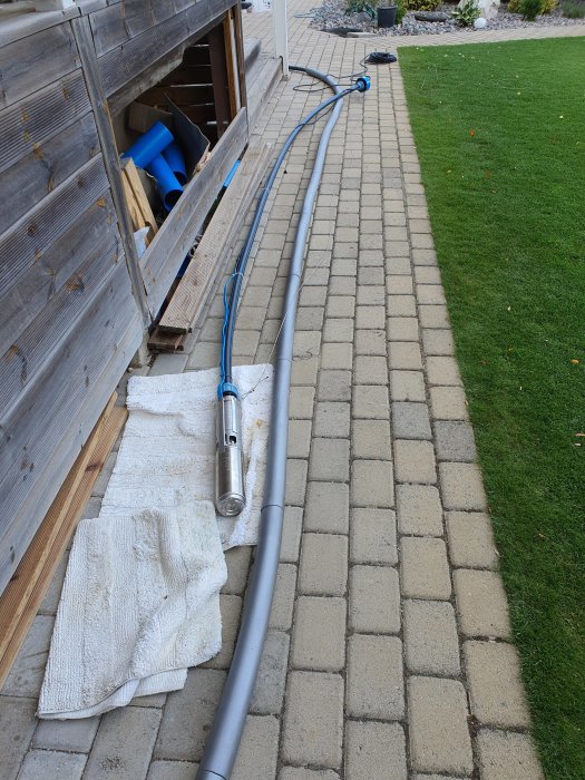PVC-rör och en djuphålspump på en handduk vid en trädgårdsstig, indikerar installation för ett kylsystem.