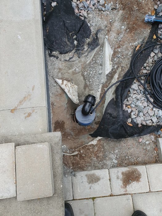 Nyligen installerad PVC-returbrunn och pump i en grävd grop vid kanten av en stenlagd yta.