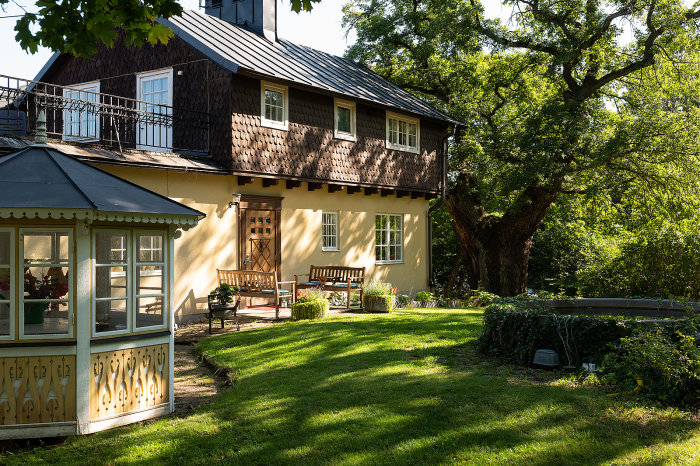 Ett hus med stalltak och en utbyggd veranda, traditionell stil, omgivet av en lummig trädgård.