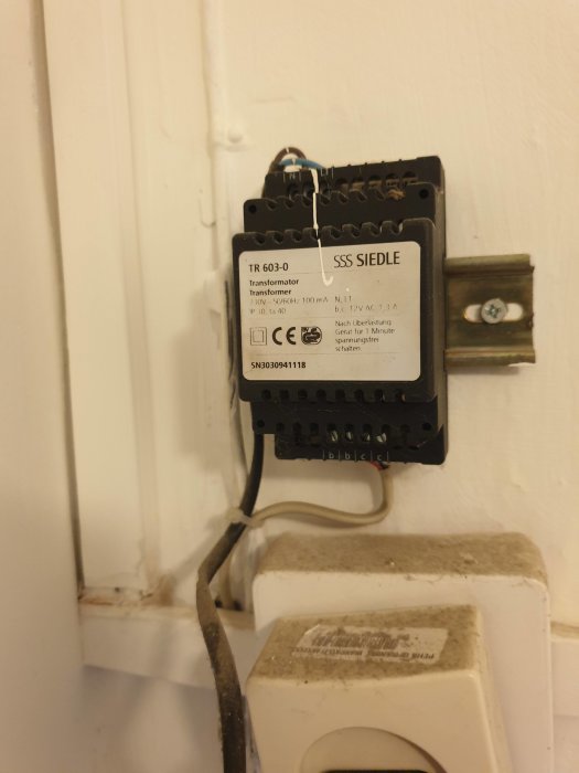 En transformator modell SSS Siedle TR 603-0 monterad på en vit vägg ovanför en smutsig, vit plastströmbrytare.