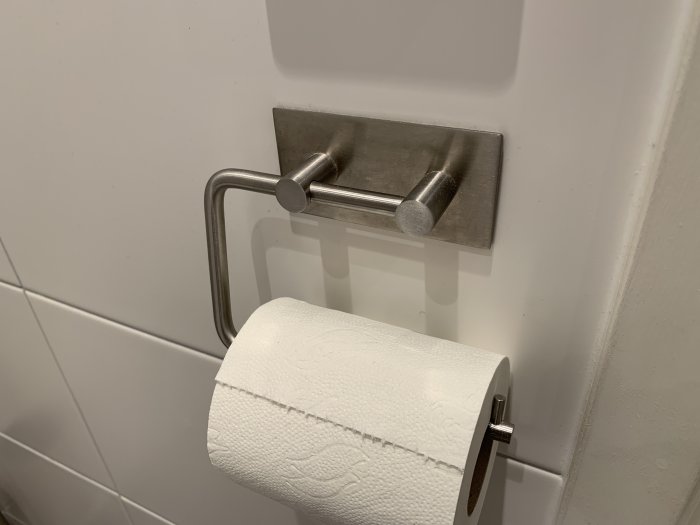 Toalettpappershållare fäst på kakelvägg med dubbelhäftande tejp, ingen borrning synlig.