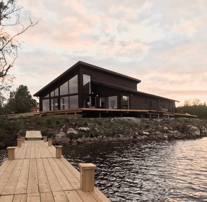 Nybyggt modernt hus med stora fönster och träbeklädnad vid vattnet, med en träbrygga som leder fram till huset.