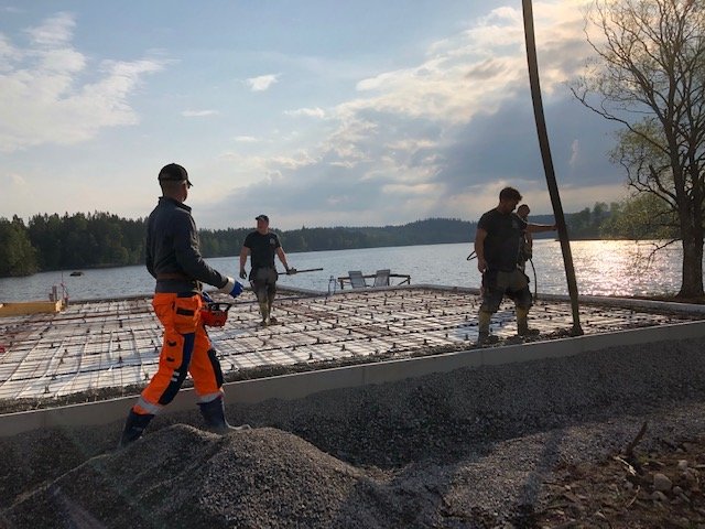 Arbetare förbereder gjutning av betongplatta vid sjö med armeringsnät och skymd solnedgång.