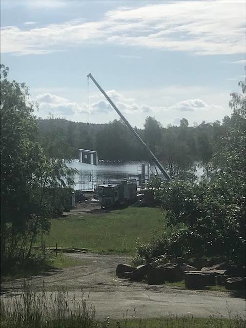 Byggarbetsplats vid vatten med mobilkran som lyfter material, gröna träd i förgrunden.