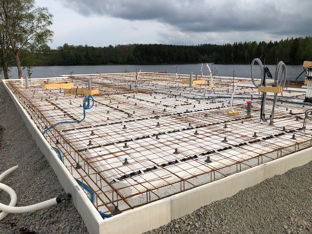 Grundläggning med armeringsjärn och gjutformar nära en sjö under molnhimmel.