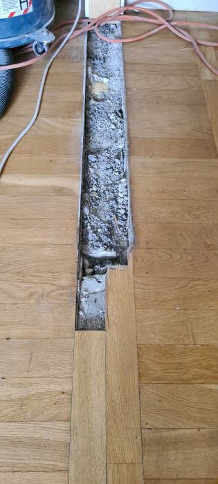 Trägolv med en nyupptagen rektangulär schakt där betong och murbruk syns efter riven vägg.